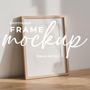 Puede incluir: Maqueta de marco de madera rectangular con un passe-partout blanco, apoyada contra una pared blanca. El marco está sobre una superficie de madera. El texto "Photoshop Frame Mockup DIN A Ratio" está superpuesto.