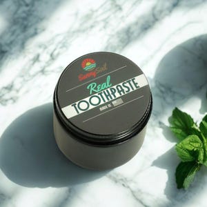 May include: A dark gray container of "Sunny Soil Real Toothpaste" with a white stripe and text. The lid features a logo of a sun and waves. The product is labeled "Made in USA". Fresh mint leaves are in the background.