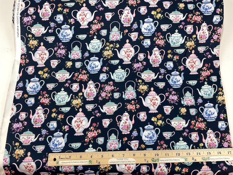 May include: Dark navy fabric with a repeating pattern of teapots, teacups, and floral arrangements in pink, blue, and yellow. The fabric has a ruler along the bottom edge. The text "DOKTIMA TEA GARDEN" is visible.