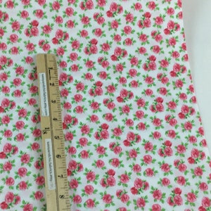 May include: White fabric covered in a pattern of small, pink roses with green leaves. A wooden ruler with black markings and text is placed on the fabric, measuring in inches.