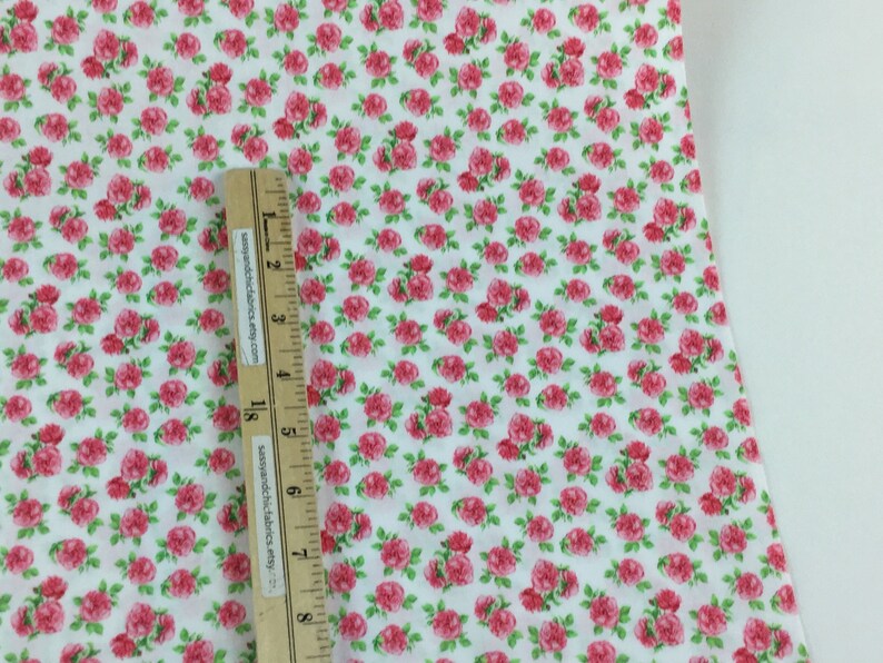 May include: White fabric covered in a pattern of small, pink roses with green leaves. A wooden ruler with black markings and text is placed on the fabric, measuring in inches.
