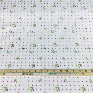 Puede incluir: Tela blanca con un patr&oacute;n repetido de puntos morados y hojas verdes con bayas moradas. Una regla de madera est&aacute; colocada sobre la tela. La tela probablemente se utiliza para proyectos de costura o artesan&iacute;a.