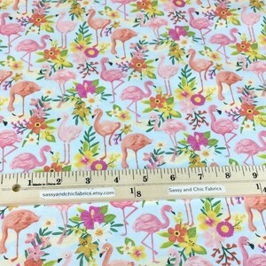 May include: Light blue fabric with a repeating pattern of pink flamingos and colorful flowers. The flowers include pink hibiscus, yellow, and orange blooms. The fabric has a ruler with inches and the text "sassyandchicfabrics.etsy.com".
