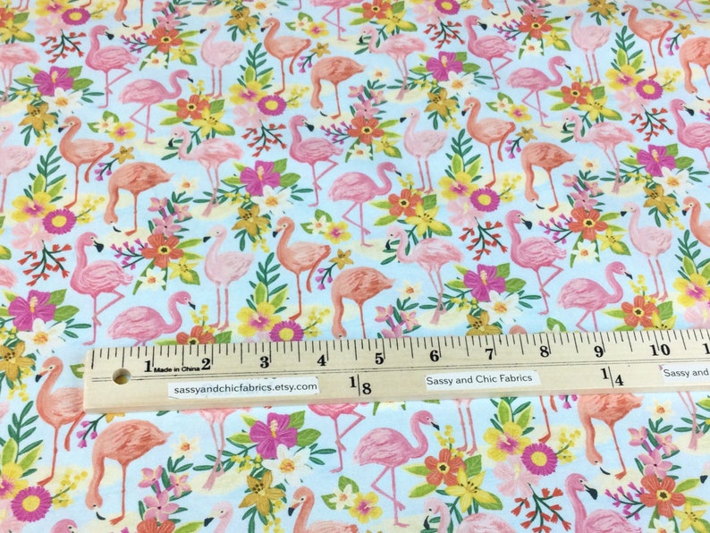 May include: Light blue fabric with a repeating pattern of pink flamingos and colorful flowers. The flowers include pink hibiscus, yellow, and orange blooms. The fabric has a ruler with inches and the text "sassyandchicfabrics.etsy.com".