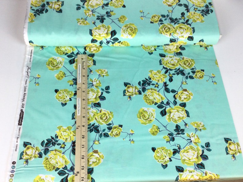 May include: A roll of fabric with a repeating floral pattern. The fabric has a light teal background with yellow roses and dark green leaves. A ruler is placed on the fabric to measure its length. The text "Love Always, AM" is visible on the edge of the fabric.
