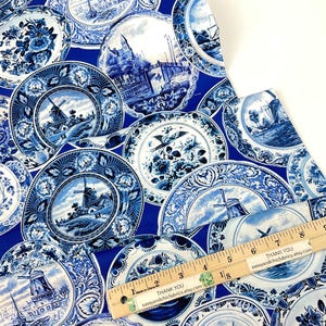 May include: Blue and white fabric featuring a repeating pattern of decorative plates. Each plate showcases a different scenic design, including windmills and floral motifs. A wooden ruler with inch measurements is visible.