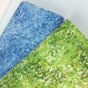 May include: Two folded fabric pieces. One is a light blue with white speckles, resembling a cloudy sky. The other is green with a floral pattern of small white flowers and touches of brown, evoking a meadow.