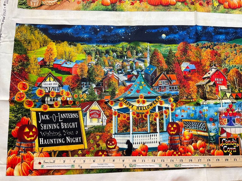 Op de afbeelding: Stoffpaneel met een Halloween-thema. De afbeelding toont een stadsgezicht met herfstbladeren, pompoenen en een prieel met een "Happy Halloween"-banner. Een bord leest "Jack-O-Lanterns Shining Bright Wishing You a Haunting Night."