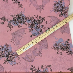 Puede incluir: Tela rosa con un patr&oacute;n repetido de murci&eacute;lagos, telara&ntilde;as y ramos florales en negro y azul claro. Una regla de madera, marcada en cent&iacute;metros, se coloca en diagonal sobre la tela. Ideal para costura o manualidades.