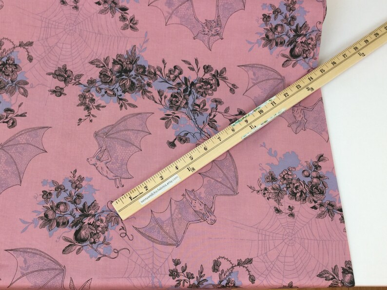 Puede incluir: Tela rosa con un patr&oacute;n repetido de murci&eacute;lagos, telara&ntilde;as y ramos florales en negro y azul claro. Una regla de madera, marcada en cent&iacute;metros, se coloca en diagonal sobre la tela. Ideal para costura o manualidades.
