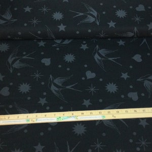 May include: Black fabric featuring a repeating pattern of grey swallows, stars, hearts, and other celestial shapes. The fabric is rolled up, showing the edge of the material. A wooden ruler is placed on the fabric.