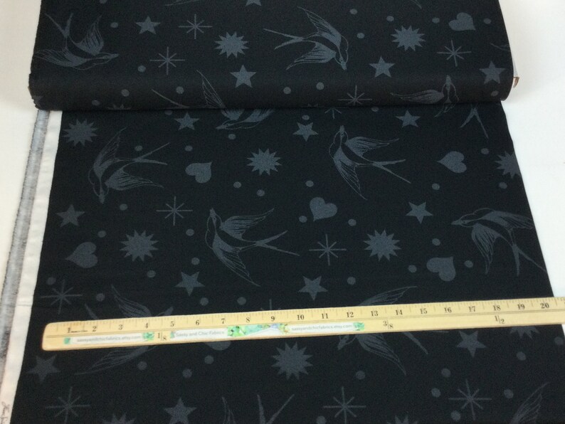 May include: Black fabric featuring a repeating pattern of grey swallows, stars, hearts, and other celestial shapes. The fabric is rolled up, showing the edge of the material. A wooden ruler is placed on the fabric.