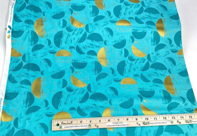 May include: Teal fabric with a gold and teal semi-circle pattern. The fabric has a textured brushstroke effect. A ruler is placed across the bottom of the fabric, measuring in inches.