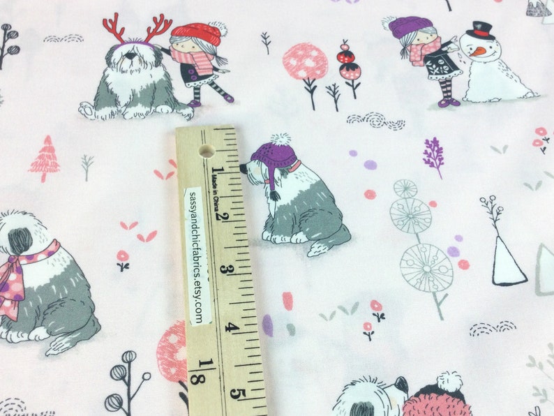 May include: Light pink fabric with a winter-themed pattern. The design features Old English Sheepdogs wearing hats and scarves, children, snowmen, and decorative trees. The fabric is decorated with small pink, purple, and gray accents.