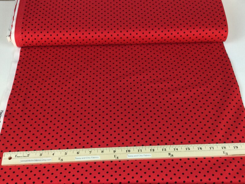 May include: Red fabric with a black polka dot pattern. A wooden ruler with inch measurements is placed on the fabric. The fabric is rolled up, showing the edge of the fabric.