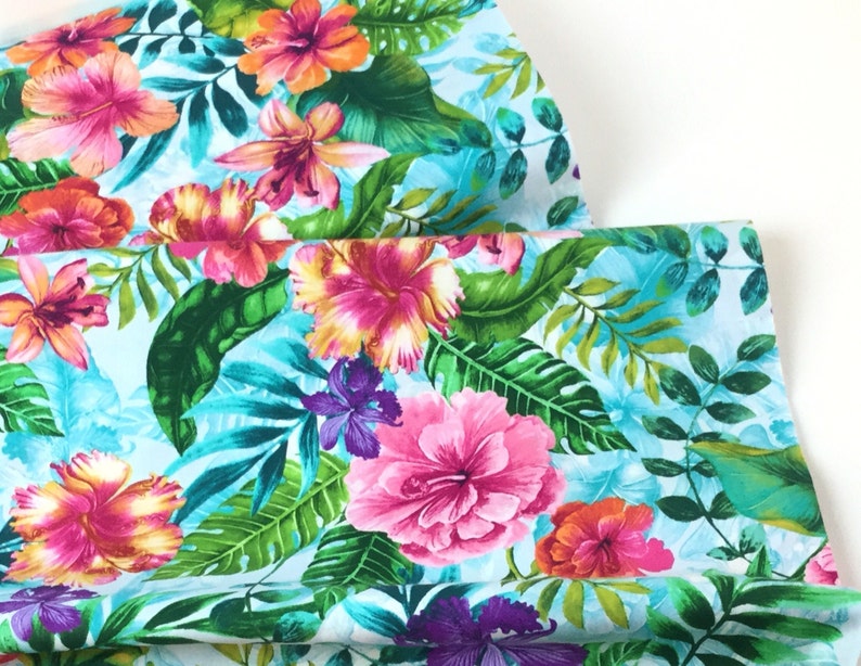 May include: Vibrant fabric featuring a tropical floral pattern. The design showcases large hibiscus flowers in shades of pink, orange, and purple, set against lush green leaves and a light blue background. The fabric is likely used for apparel or home decor.