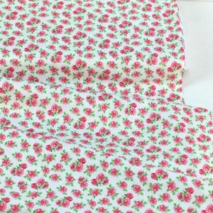 May include: White fabric covered in a pattern of small pink roses with green leaves. The fabric is folded over, showing the pattern repeat. This floral fabric is suitable for sewing projects, quilting, and crafting.