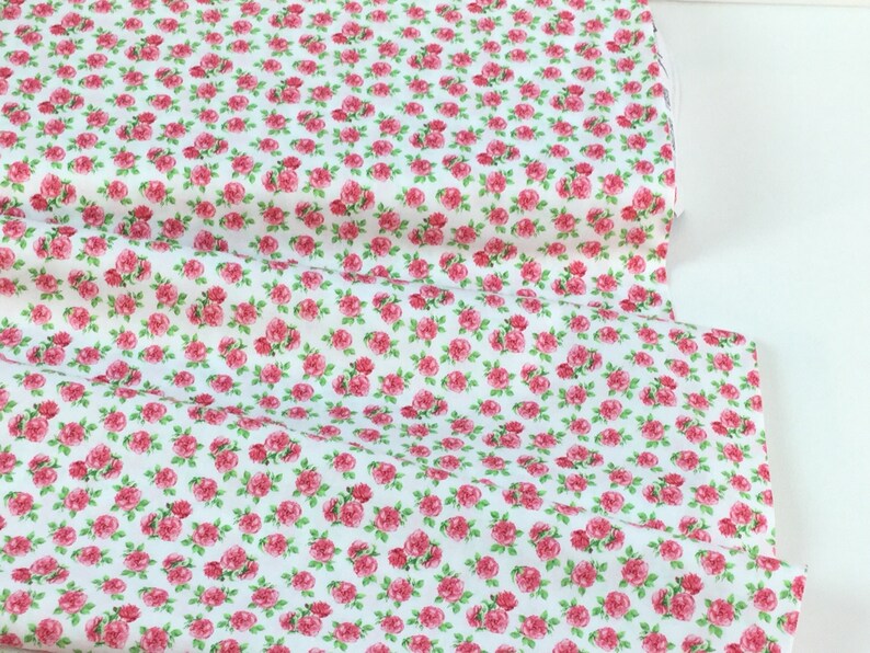 May include: White fabric covered in a pattern of small pink roses with green leaves. The fabric is folded over, showing the pattern repeat. This floral fabric is suitable for sewing projects, quilting, and crafting.