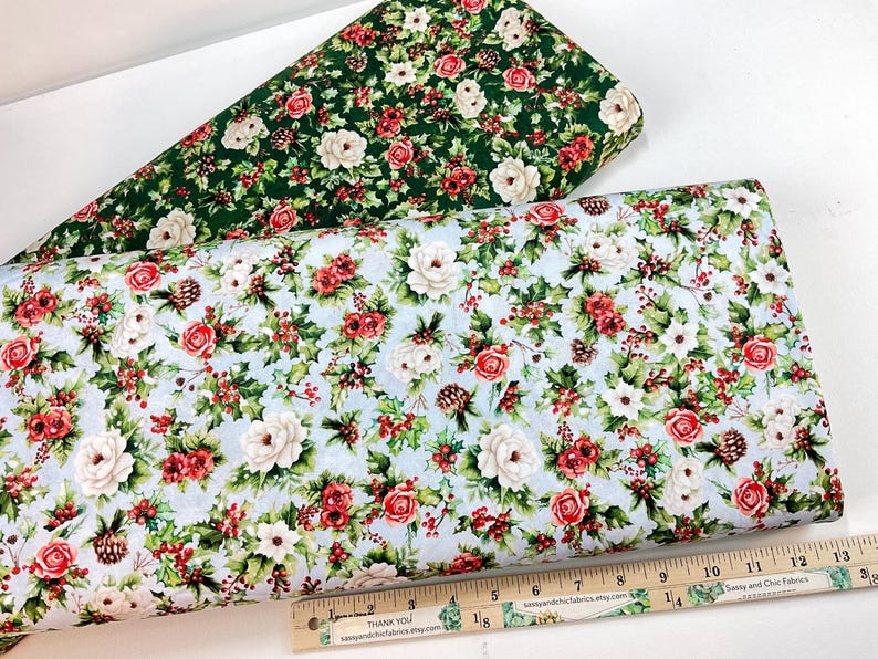 K&ouml;nnte beinhalten: Zwei Stoffrollen mit floralen Mustern. Eine Rolle hat einen gr&uuml;nen Hintergrund mit wei&szlig;en und roten Blumen. Die andere hat einen hellblauen Hintergrund mit wei&szlig;en und roten Blumen, Tannenzapfen und Beeren. Ein Lineal befindet sich unten.