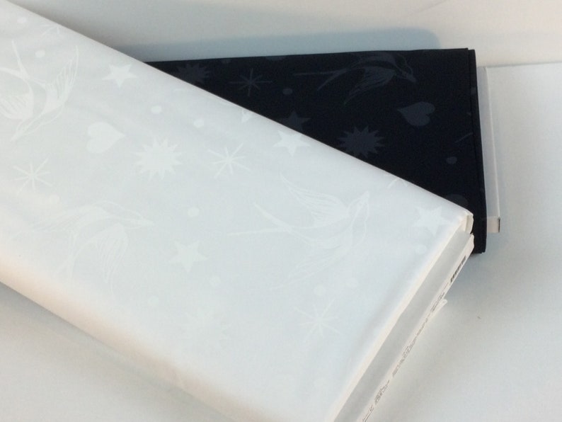 May include: Two folded fabric samples, one white and one black, are stacked. The white fabric displays a repeating pattern of birds, stars, and hearts. The black fabric has a similar design. Both fabrics appear to be cotton or a similar material.