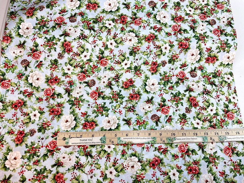K&ouml;nnte beinhalten: Blumenstoff mit hellblauem Hintergrund, mit einem Muster aus roten und wei&szlig;en Rosen, gr&uuml;nen Bl&auml;ttern und kleinen Tannenzapfen. Ein Holzlineal mit Zollma&szlig;en ist sichtbar.