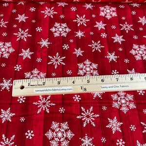 Pu&ograve; includere: Tessuto rosso con un motivo a fiocchi di neve bianchi. Il tessuto ha un aspetto venato del legno. Un righello di legno con misure in centimetri &egrave; in basso. Il testo "Sassy and Chic Fabrics" &egrave; visibile.