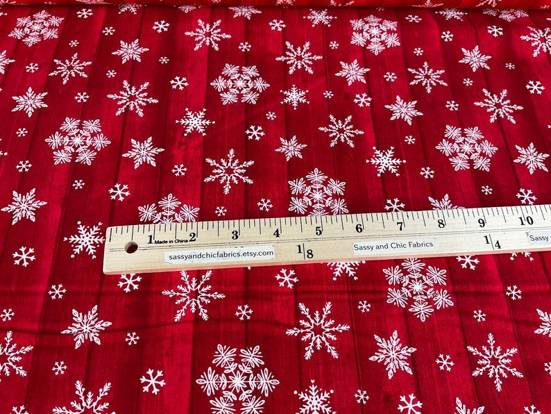 Pu&ograve; includere: Tessuto rosso con un motivo a fiocchi di neve bianchi. Il tessuto ha un aspetto venato del legno. Un righello di legno con misure in centimetri &egrave; in basso. Il testo "Sassy and Chic Fabrics" &egrave; visibile.