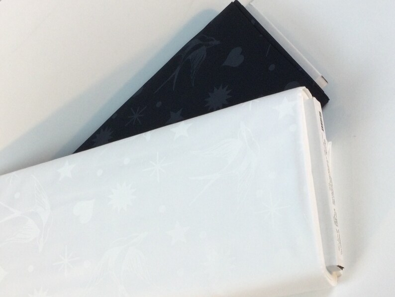 May include: Two rolls of fabric, one white and one black. The white fabric features a pattern of stars, birds, and hearts. The black fabric has a similar design. The rolls are stacked on top of each other.