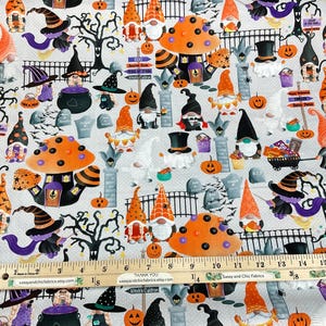May include: Halloween-themed fabric featuring gnomes, pumpkins, witch hats, and tombstones on a light gray background. The design includes orange and black mushrooms, trees, and signs. A ruler shows measurements in inches.