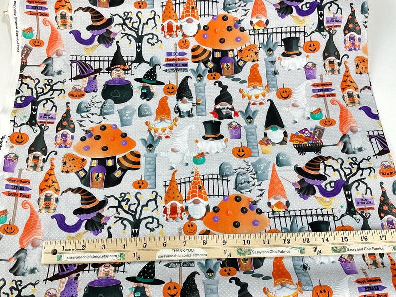 May include: Halloween-themed fabric featuring gnomes, pumpkins, witch hats, and tombstones on a light gray background. The design includes orange and black mushrooms, trees, and signs. A ruler shows measurements in inches.