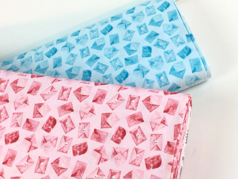 May include: Two fabric bolts, one light blue and one pink, are patterned with watercolor-style envelopes. The envelopes have a button closure and the number "3" printed on them. The fabric is likely cotton or a similar material.
