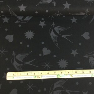 May include: Black fabric featuring a repeating pattern of grey swallows, stars, hearts, and dots. The fabric is rolled up, displaying the design across the surface. A ruler is placed on the fabric for scale.