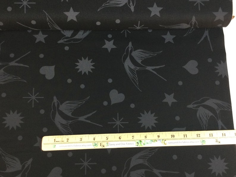 May include: Black fabric featuring a repeating pattern of grey swallows, stars, hearts, and dots. The fabric is rolled up, displaying the design across the surface. A ruler is placed on the fabric for scale.