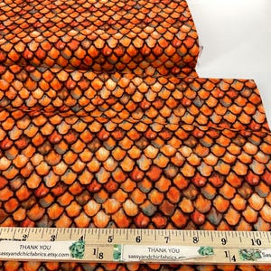 May include: Orange and black fabric with a scale pattern. The fabric is printed with a repeating design of overlapping, textured scales in shades of orange, brown, and black. A wooden ruler with inch measurements is in the image.