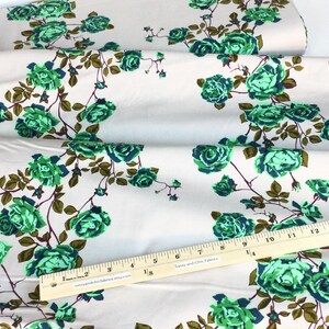 May include: Fabric with a repeating floral pattern of green roses and brown stems on a cream background. A wooden ruler with inch markings is placed across the fabric. The fabric is folded in layers.