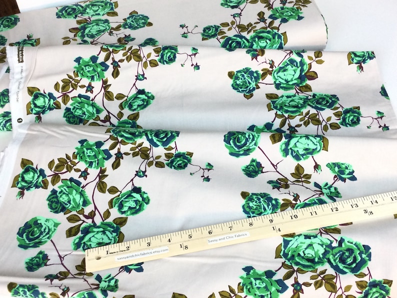 May include: Fabric with a repeating floral pattern of green roses and brown stems on a cream background. A wooden ruler with inch markings is placed across the fabric. The fabric is folded in layers.