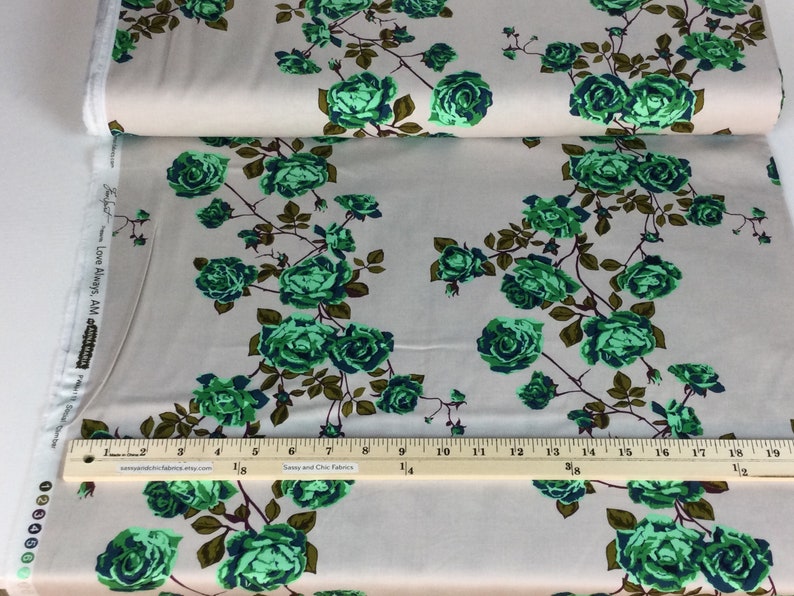 May include: Fabric with a floral pattern of green roses and brown stems on a light gray background. The fabric is folded over, showing the edge of the roll. A ruler is placed on the fabric for measurement.