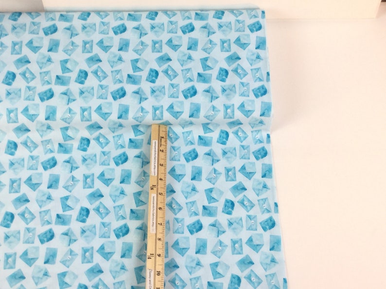 May include: Light blue fabric with a repeating pattern of watercolor-style envelopes. The envelopes are in shades of blue and have a button detail. A ruler is placed on the fabric, showing inches.