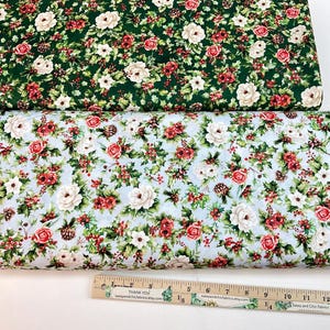 K&ouml;nnte beinhalten: Zwei Stoffrollen mit floralen Mustern. Eine hat einen dunkelgr&uuml;nen Hintergrund mit wei&szlig;en Blumen, roten Beeren und gr&uuml;nen Bl&auml;ttern. Die andere hat einen hellblauen Hintergrund mit roten Rosen, wei&szlig;en Blumen und gr&uuml;nen Bl&auml;ttern. Unten ist ein Lineal.