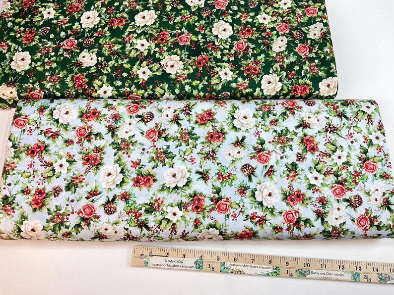 K&ouml;nnte beinhalten: Zwei Stoffrollen mit floralen Mustern. Eine hat einen dunkelgr&uuml;nen Hintergrund mit wei&szlig;en Blumen, roten Beeren und gr&uuml;nen Bl&auml;ttern. Die andere hat einen hellblauen Hintergrund mit roten Rosen, wei&szlig;en Blumen und gr&uuml;nen Bl&auml;ttern. Unten ist ein Lineal.