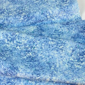 May include: A close-up of a fabric with a blue and white floral pattern. The fabric has a soft, textured appearance with a watercolor effect. The color palette includes shades of blue and white.