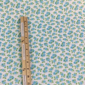 May include: White fabric with a pattern of small blue roses and green leaves. A wooden ruler with text is placed on the fabric, measuring in inches. The fabric has a floral design.