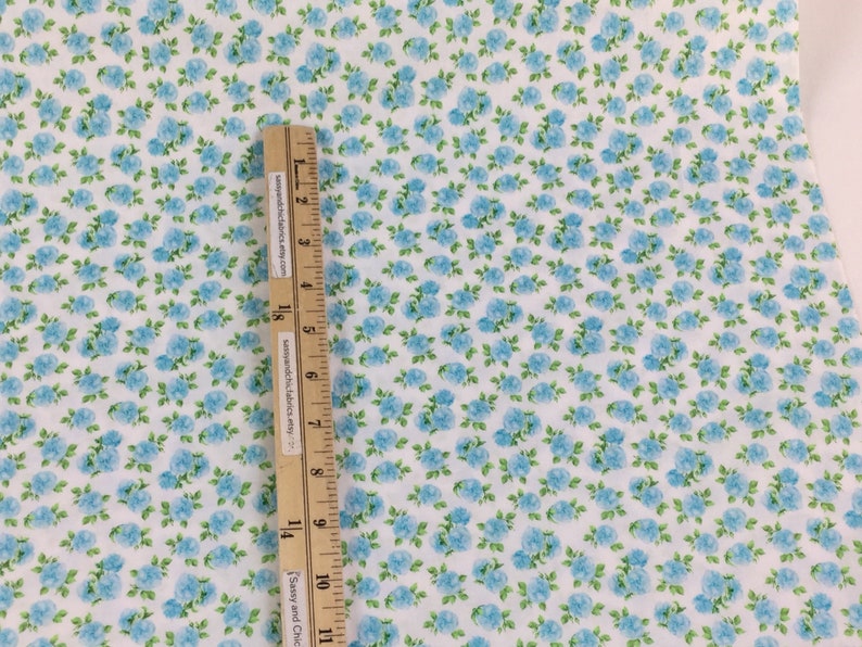 May include: White fabric with a pattern of small blue roses and green leaves. A wooden ruler with text is placed on the fabric, measuring in inches. The fabric has a floral design.
