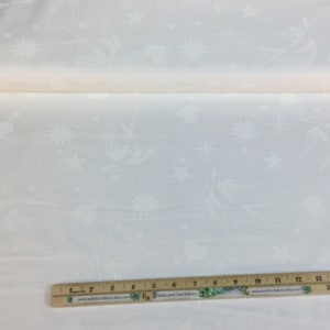 May include: A roll of white fabric featuring a repeating pattern of birds, stars, hearts, and sunbursts. The fabric is a light colour and appears to be a cotton or linen blend. A wooden ruler with inch markings is at the bottom.
