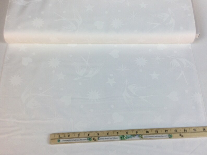 May include: A roll of white fabric featuring a repeating pattern of birds, stars, hearts, and sunbursts. The fabric is a light colour and appears to be a cotton or linen blend. A wooden ruler with inch markings is at the bottom.