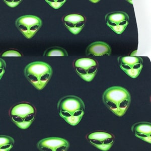May include: Black fabric featuring a repeating pattern of green alien faces. The aliens have large black eyes and are set against a dark background. The fabric is likely used for apparel or home decor.