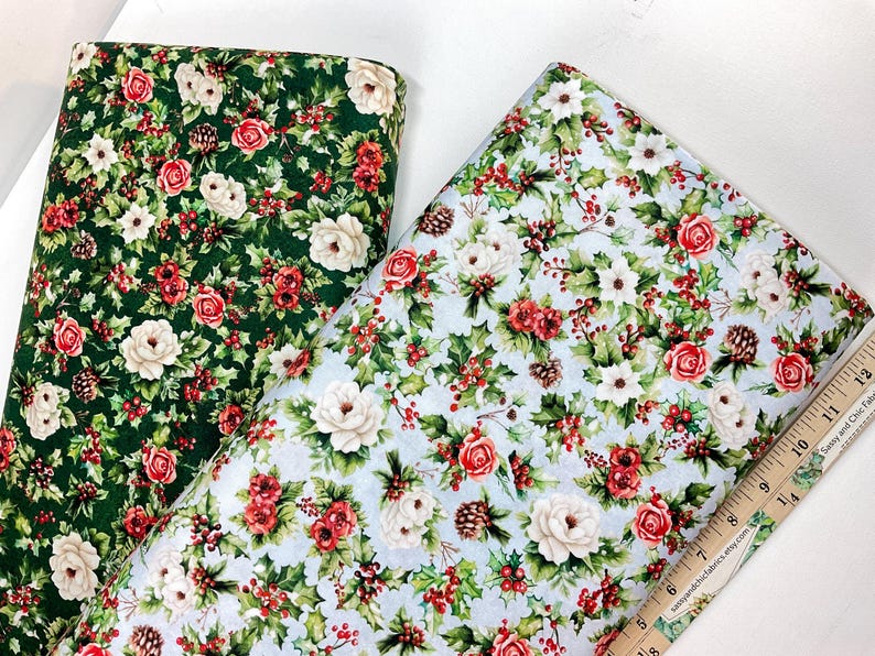 K&ouml;nnte beinhalten: Zwei Stoffrollen mit floralen Mustern. Eine hat einen dunkelgr&uuml;nen Hintergrund mit roten und wei&szlig;en Rosen, Stechpalmen und Tannenzapfen. Die andere hat einen hellblauen Hintergrund mit dem gleichen floralen Design. Rechts ist ein Holzlineal.