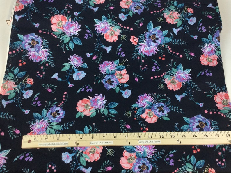May include: Dark navy fabric with a repeating floral pattern. The design features clusters of pink, purple, and blue flowers with green foliage. A ruler with inch measurements is visible.