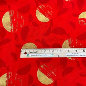 May include: Red fabric with a repeating pattern of gold half-circles and full circles. The gold elements have a brushstroke texture. A ruler is placed across the fabric, measuring in inches.