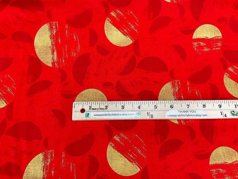 May include: Red fabric with a repeating pattern of gold half-circles and full circles. The gold elements have a brushstroke texture. A ruler is placed across the fabric, measuring in inches.